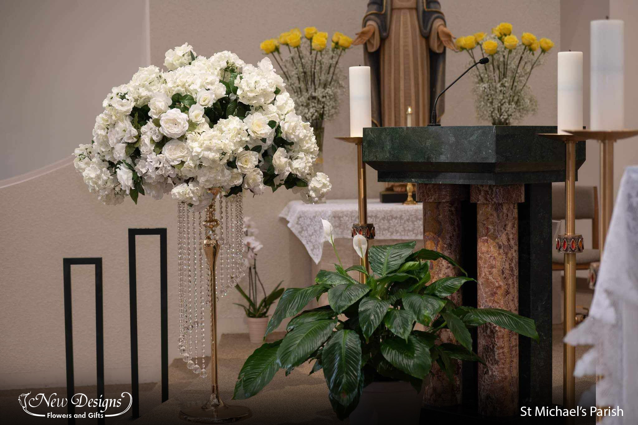 A tall arrangement sits at the front of a church. A large compact Floral arrangement of White hydrangeas and white roses sits a top a tall gold stand, with draping crystals descending in a spiral form.